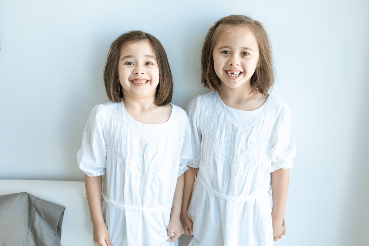 Two young girls happily smiling in matching white dresses, captured indoors with a bright ambiance.